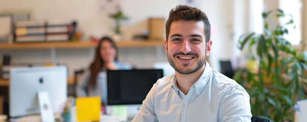 Wall Mural - Smiling young businessman in a modern office, wearing a light blue shirt. Background shows a coworking space with a blurred female colleague and office supplies.