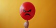 © BoOm - Hand holding a red angry face balloon against a vibrant yellow background, representing emotion, frustration, and negative feelings.