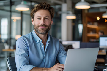 Portrait of a successful and confident young man in a blue shirt sitting in the office at a desk, working on a laptop and smiling at the camera
