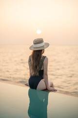 Wall Mural - Woman in black swimsuit relaxing by infinity pool with ocean view during sunset.