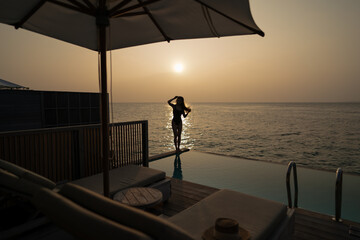 Wall Mural - Woman in black swimsuit relaxing by infinity pool with ocean view during sunset.