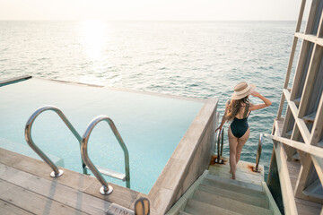 Wall Mural - Woman in black swimsuit relaxing by infinity pool with ocean view.