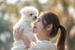 © Sourav Mittal - Asian woman playing with white bichon frise puppy in the park, holding and lifting the dog up to her face, outdoor portrait of a happy pet owner with a small fluffy pure animal, side view, blurred bac