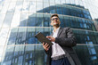 © muse studio - A confident businessman in a suit holds a tablet, standing against a backdrop of modern glass buildings.