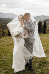 Wall Mural - A bride and groom are standing in a field with a white veil over the bride. The bride is holding a bouquet of flowers and the groom is wearing a suit. The scene is romantic and intimate