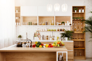 A modern kitchen island with fresh fruit and vegetables on display. The island is made of light wood and has a white countertop. There is a kettle on the stovetop and other kitchen supplies in the