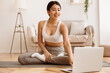 © Prostock-studio - A young woman in a white sports bra and grey pants is sitting on a yoga mat in a living room, following along with a yoga video on her laptop.