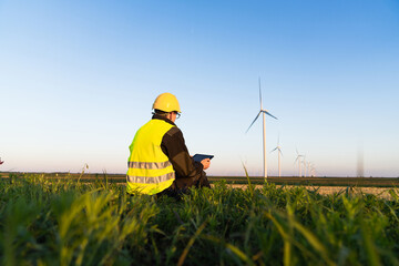 Canvas Print - Worker with digital tablet sits on the grass and looks at wind turbines.