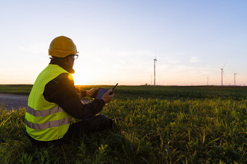 Aufkleber - Worker with digital tablet sits on the grass and looks at wind turbines.