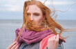 © ullision - A woman with red hair stands by the sea on a windy day, hair is blowing across her face, partially obscuring it, wears a purple scarf and a pink jacket, looking directly at camera