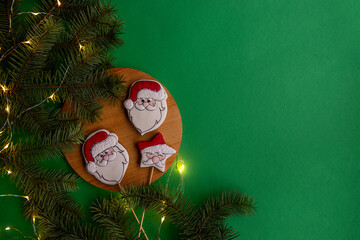 Three Santa Claus-shaped cookies on a wooden board, surrounded by pine branches and festive lights on a green background.