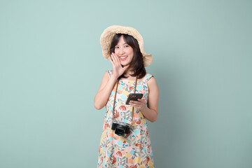 Wall Mural - Travel and vacation promotion concept. Happy Asian woman wearing casual dress and hat with camera and mobile phone while shouting to free copy space isolated on pastel green background.