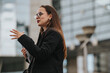 © qunica.com - Confident businesswoman wearing glasses, gesturing while talking outside a modern office building. Professional, focused, and determined in a corporate environment.