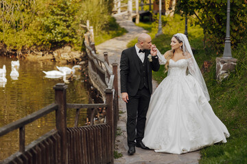 Wall Mural - A bride and groom walk along a path near a pond. The bride is wearing a white dress and the groom is wearing a black suit. There are several ducks in the pond