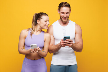 Wall Mural - Young fun strong fitness trainer instructor sporty two man woman wear blue clothes spend time in home gym hold use mobile cell phone app isolated on plain yellow background. Workout sport fit concept.