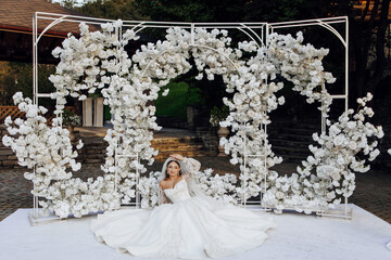 Wall Mural - A woman is sitting in front of a white archway with flowers. The woman is wearing a white dress and is posing for the camera. The scene is elegant and romantic