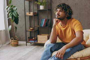 Wall Mural - Young minded pensive thoughtful Indian man he wear orange casual clothes prop up chin sits in armchair stay at home hotel flat rest relax spend free spare time in living room indoor. Lounge concept.
