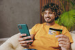 © ViDi Studio - Young Indian man wearing casual clothes hold mobile cell phone credit bank card shopping online sits in armchair stay at home hotel flat rest relax spend free spare time in living room Lounge concept