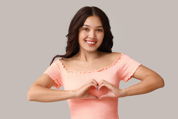 Wall Mural - Happy young Asian woman showing heart gesture on grey background. Valentine's Day celebration