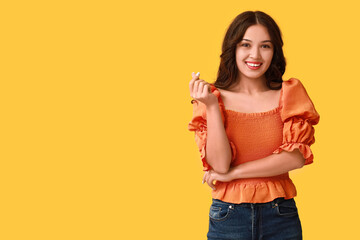 Wall Mural - Happy young Asian woman showing heart gesture on yellow background. Valentine's Day celebration