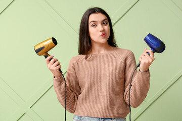 Wall Mural - Beautiful young thoughtful woman with hair dryers on green background