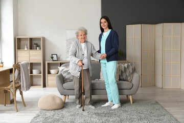 Wall Mural - Young caregiver helping senior woman with stick to walk at home