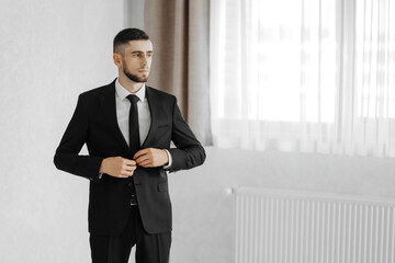 A man in a suit and tie is standing in front of a window. He is adjusting his tie and looking out the window. Concept of formality and professionalism, as the man is dressed in a suit and tie