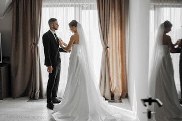 Wall Mural - A bride and groom are getting ready for their wedding. The bride is wearing a white dress and the groom is wearing a suit. They are standing in front of a window with curtains