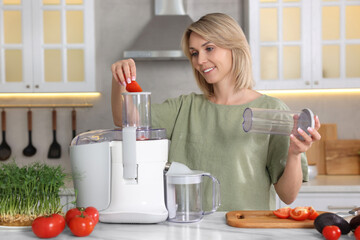 Wall Mural - Smiling woman putting fresh tomato into juicer at white marble table in kitchen