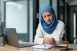 © crizzystudio - Modern office Muslim businesswoman wearing hijab sits and works with laptop online for a long time from fatigue and arm pain, meditate and stretch to relax. Office syndrome concept.