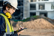 © Summer Paradive - Asian male site engineer or building inspector man with green reflective vest, safety helmet, goggles and earmuffs using digital tablet inspecting working process at construction site.
