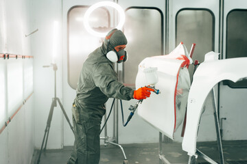 Auto mechanic worker wearing protective workwear spraying white paint on car part at workshop