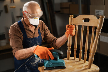 An elderly man in orange gloves, blue apron, protective mask and glasses sands an old wooden chair with a sanding machine in a workshop.