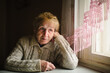 © De Visu - An elderly woman in a sweater sits by a window, gazing outside with a serene expression, capturing a moment of peaceful reflection.