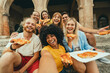 © Davide Angelini - Multicultural smiling friends eating pizza sitting outside - Cheerful young people taking selfie photo with smartphone outdoor - Happy life style concept with guys and girls hanging out on a sunny day
