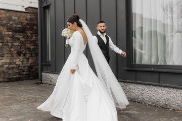 Wall Mural - A bride and groom are walking down a street, the bride is holding a bouquet and the groom is wearing a vest