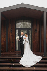 Wall Mural - A bride and groom stand on a wooden staircase in front of a house. The bride is wearing a white dress and the groom is wearing a black suit. They are holding a bouquet and a vase, respectively