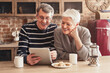 © Prostock-studio - A happy senior couple are sitting in their kitchen, looking at a tablet computer. The woman has a hand on her chin and is laughing, while the man is holding the tablet and smiling