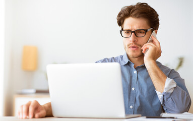 Leinwandbilder - A man in a blue shirt and glasses sits at a desk, talking on his phone while looking at his laptop.