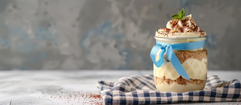 An unusual and appetizing tiramisu presentation in a jar with a blue ribbon on a checkered napkin against a concrete backdrop, ideal for copy space image.