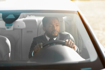 Wall Mural - Black man in a suit driving a car. The man is focused on the road and is behind the wheel of the vehicle. The sun is shining through the windshield, and the car appears to be on a sunny day.