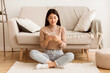 © Prostock-studio - A young woman in a beige sweater and blue jeans sits on the floor in front of a couch, using her smartphone.