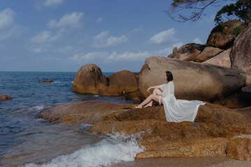 Woman in white robe sits on rock by calm blue ocean, legs outstretched towards water. White clouds in sky, seems content in peaceful view