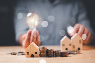 Person is holding light bulb and stack of coins in front of two wooden houses. Scene suggests that person is trying to come up with idea or solution to problem, loan, estate, real, growing, success