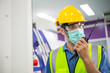© winnievinzence - Male worker wearing medical mask talking on walkie-talkie with colleague to control work in front of technology machine at industrial plant factory. copy space