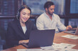 © NT_studio - Professional young asian businesswoman wearing headset working on laptop in modern office with colleague in background, business woman wearing headphone for customer service, business concept.