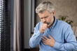 © Liubomir - Elderly man with grey hair and beard coughing and holding chest with painful expression. Health concept of illness, bronchitis, lung disease, or flu. Indoors with window and blurred background.
