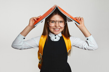 Wall Mural - Positive little blonde girl wearing eyeglasses and yellow backpack holding open copybook on head while smiling broadly against white background