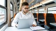 © lililia - A young man wearing glasses works on his laptop computer while riding a train. He is sitting in a seat facing forward, with a window on his left and other seats behind him