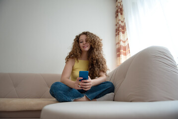 Portrait of a beautiful cheerful woman with curly hair sitting on a sofa at home and using a mobile phone